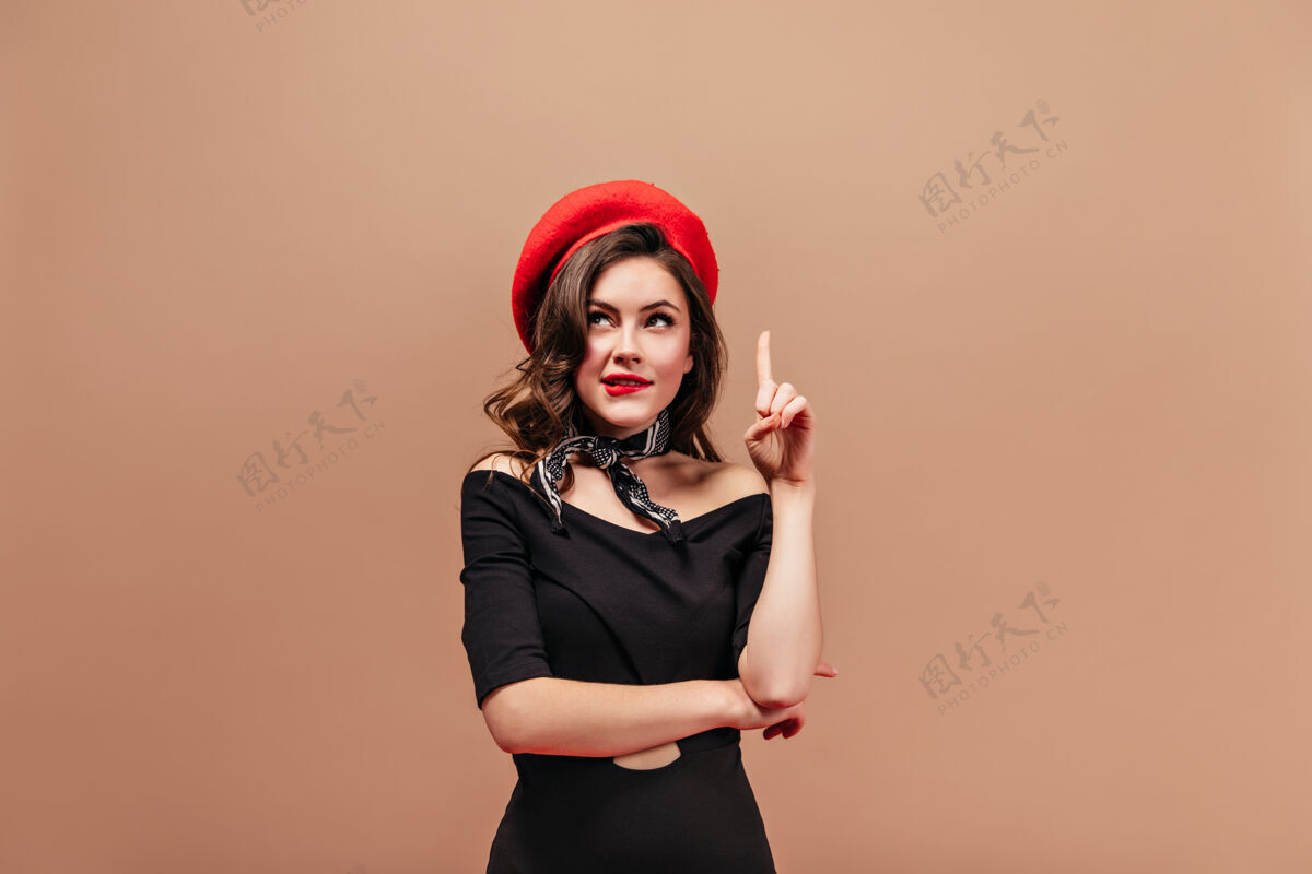小卷曲的红唇女士若有所思地抬起头来 食指向上戴着明亮贝雷帽和黑色连衣裙的女人的肖像红色长米色背景
