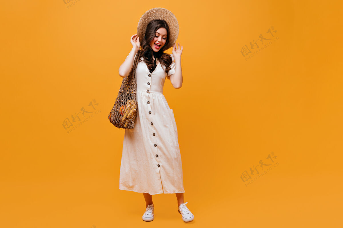 波浪发穿着棉质迷笛裙和运动鞋的女士戴着宽边草帽深色皮肤的女士拿着橙色背景的购物袋摆姿势黑发女性成人