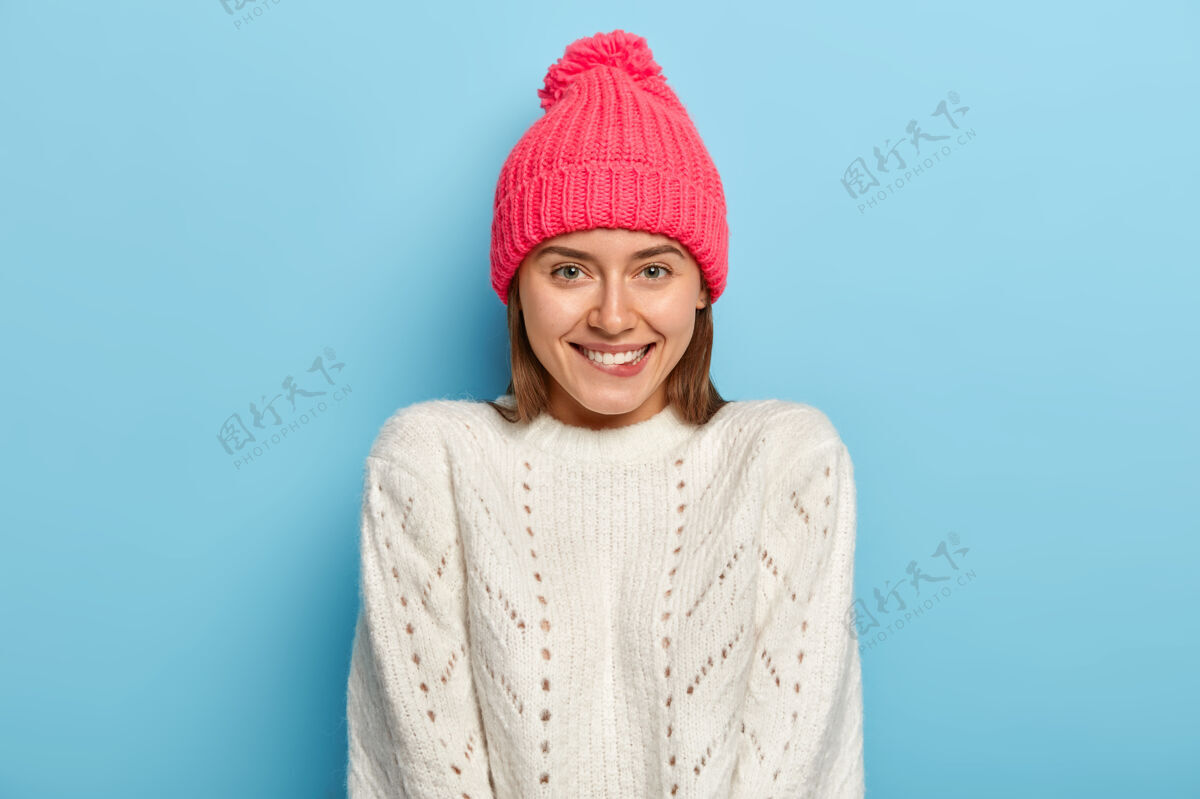 年轻娇美的女孩 神采奕奕 咬着嘴唇 高兴地看着镜头 戴着粉色的帽子 穿着白色的毛衣 隔着蓝色的墙壁 感觉在家的舒适姿势穿着快乐
