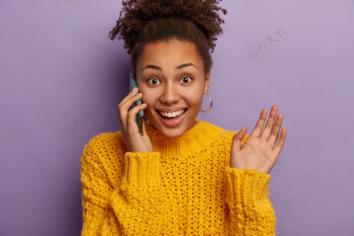 年轻快乐的卷发女青年讲电话 高兴听到好消息 在交谈中做手势 举起手掌 戴耳环和黄色毛衣 喜欢随意交谈 孤立在紫色的背景下肖像快乐卷发