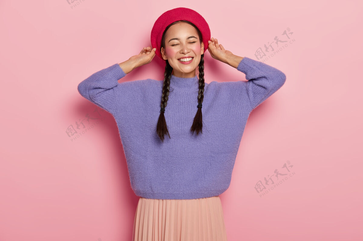 毛衣快乐的亚洲女人抚摸贝雷帽 愉快地微笑 露出白色的牙齿 留着两条辫子 穿着紫色的套头衫和裙子 表达愉快的心情 隔着粉色的墙满足高兴亚洲人