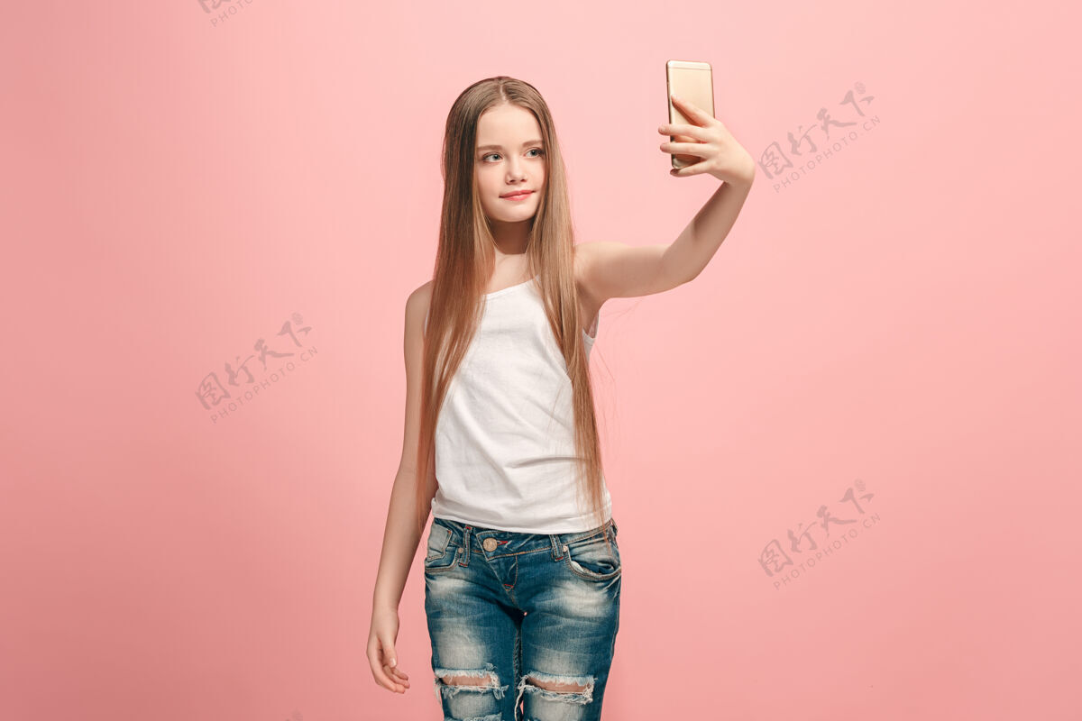 满意快乐的少女站在粉红色的墙上 微笑着 用手机自拍人类的情感 面部表情概念正面视图电话站立女孩