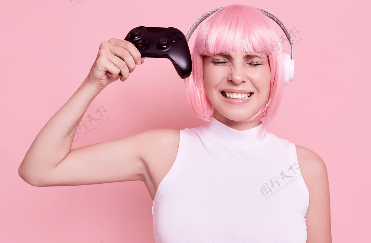 微笑美丽的粉红色头发的女人在工作室玩游戏手柄的肖像耳机疯狂玩家