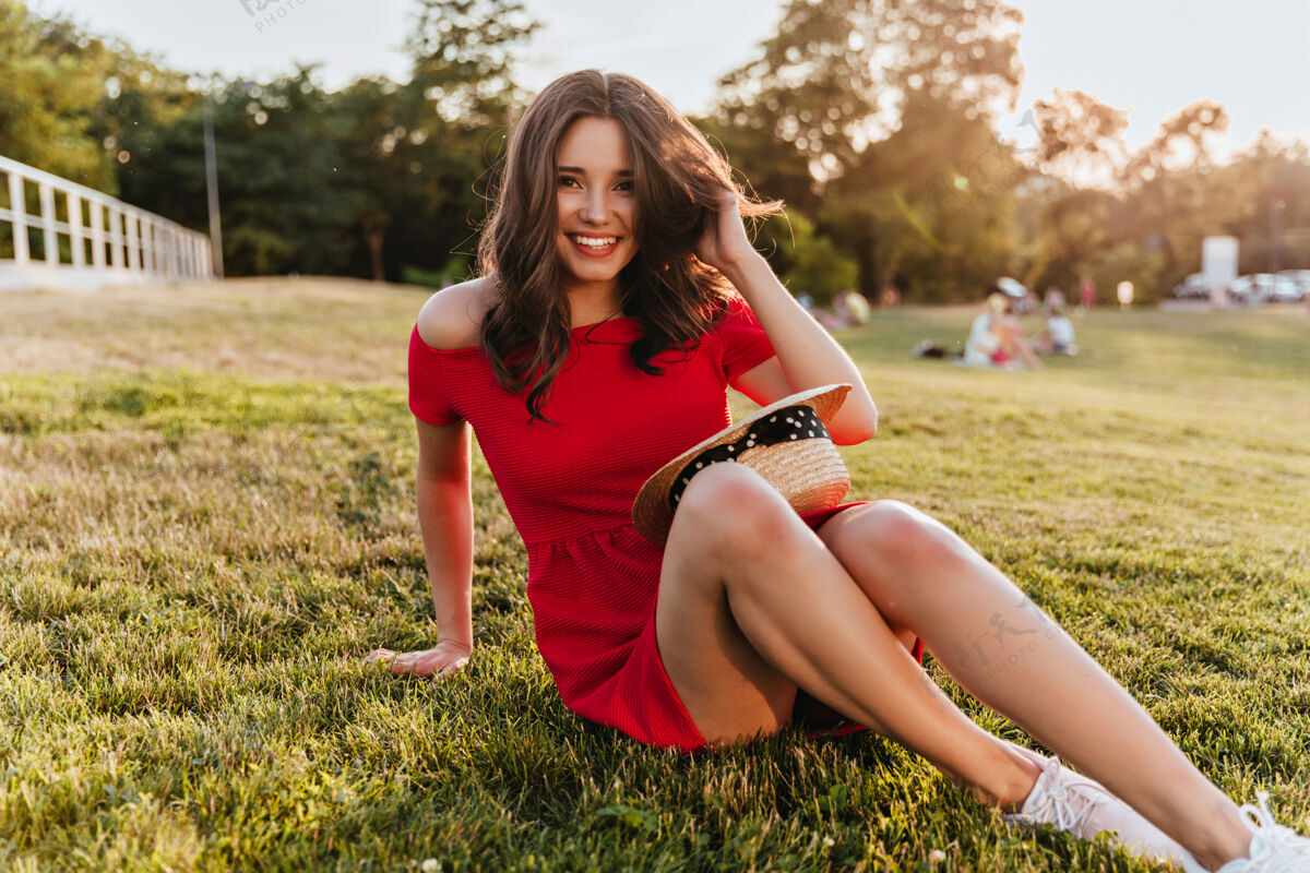 减肥美丽的女孩 羞涩的微笑 坐在公园的地上阳光明媚的日子里 一个穿着红裙子 幽默的黑发女人在草地上摆姿势年轻放松户外