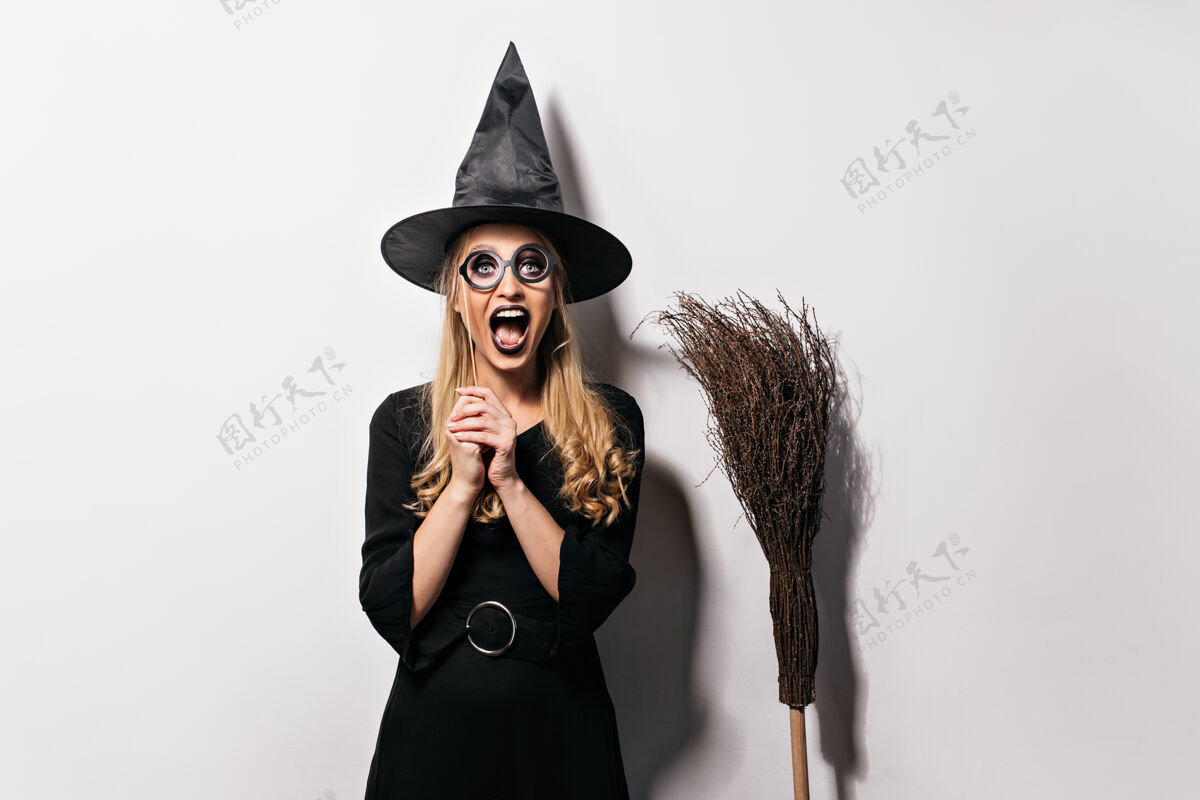 肖像欢笑的金发女孩在万圣节的化妆舞会上幽默的女巫戴着黑帽子摆姿势享受巫术化妆舞会