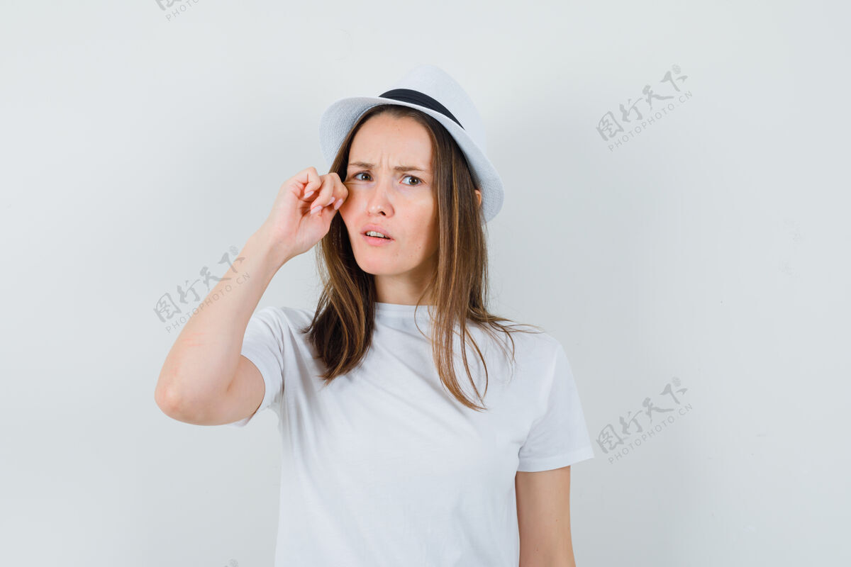 快乐年轻女孩拉着她的脸颊在白色t恤 帽子和困惑的样子前视图孩子可爱快乐