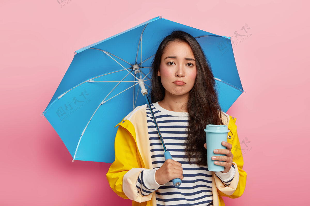 年轻心烦意乱的韩国女孩雨伞下散步 喝外卖咖啡 穿着防水雨衣美女人饮酒