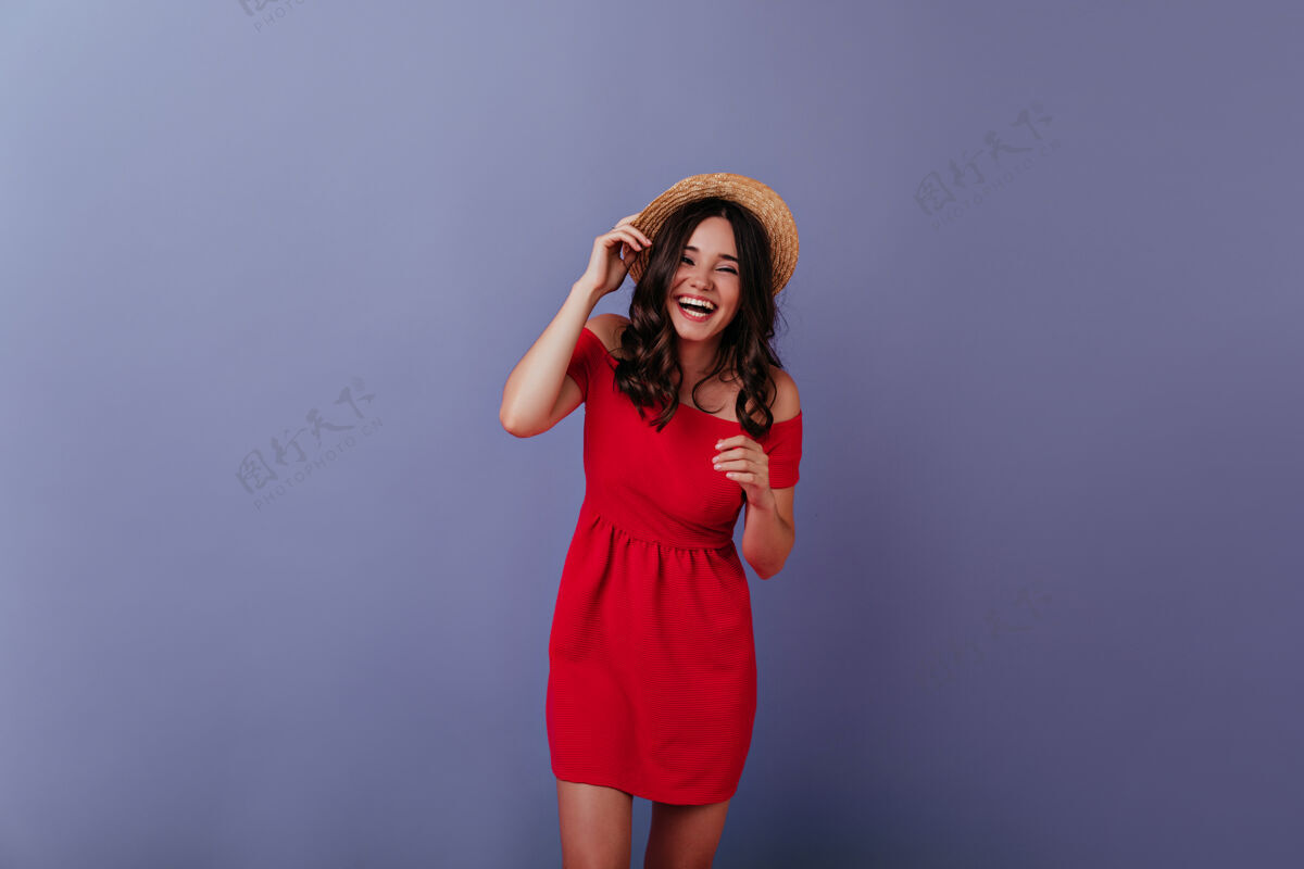 年轻可爱的棕色头发的女孩在紫色的墙上欢笑室内照片快乐的棕发女士穿着红色的裙子和夏天的帽子服装室内服饰