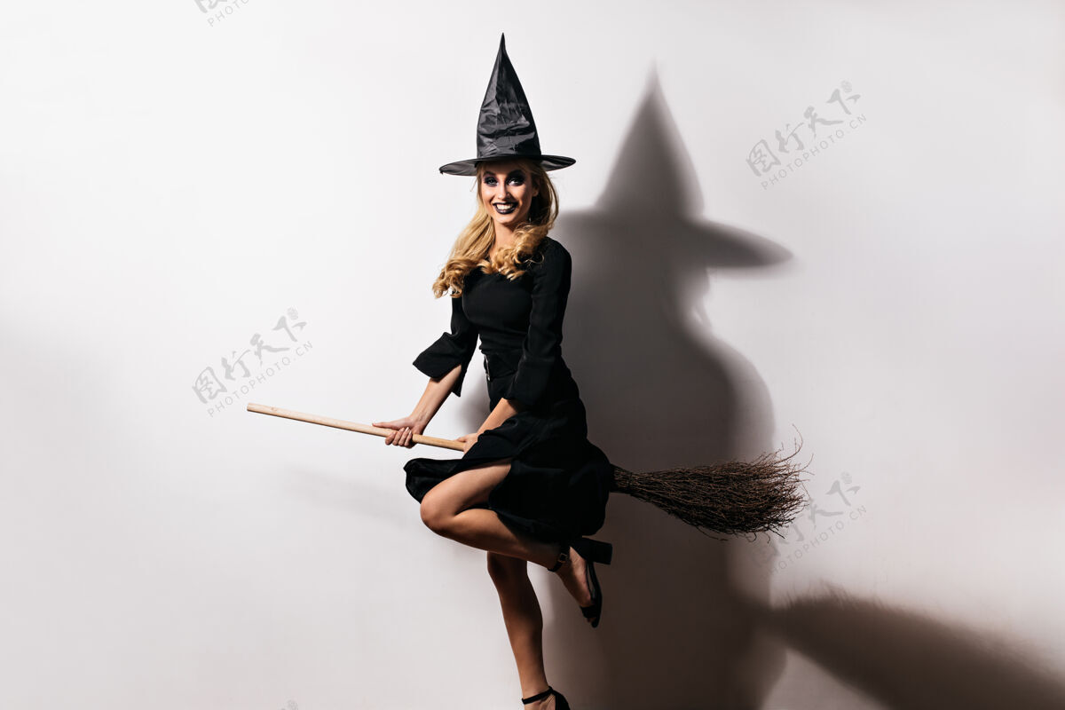 扫帚戴着女巫帽的欣喜若狂的年轻女子在狂欢节中玩得很开心优雅的白人女孩坐在魔法扫帚上的室内照片享受万圣节邪恶