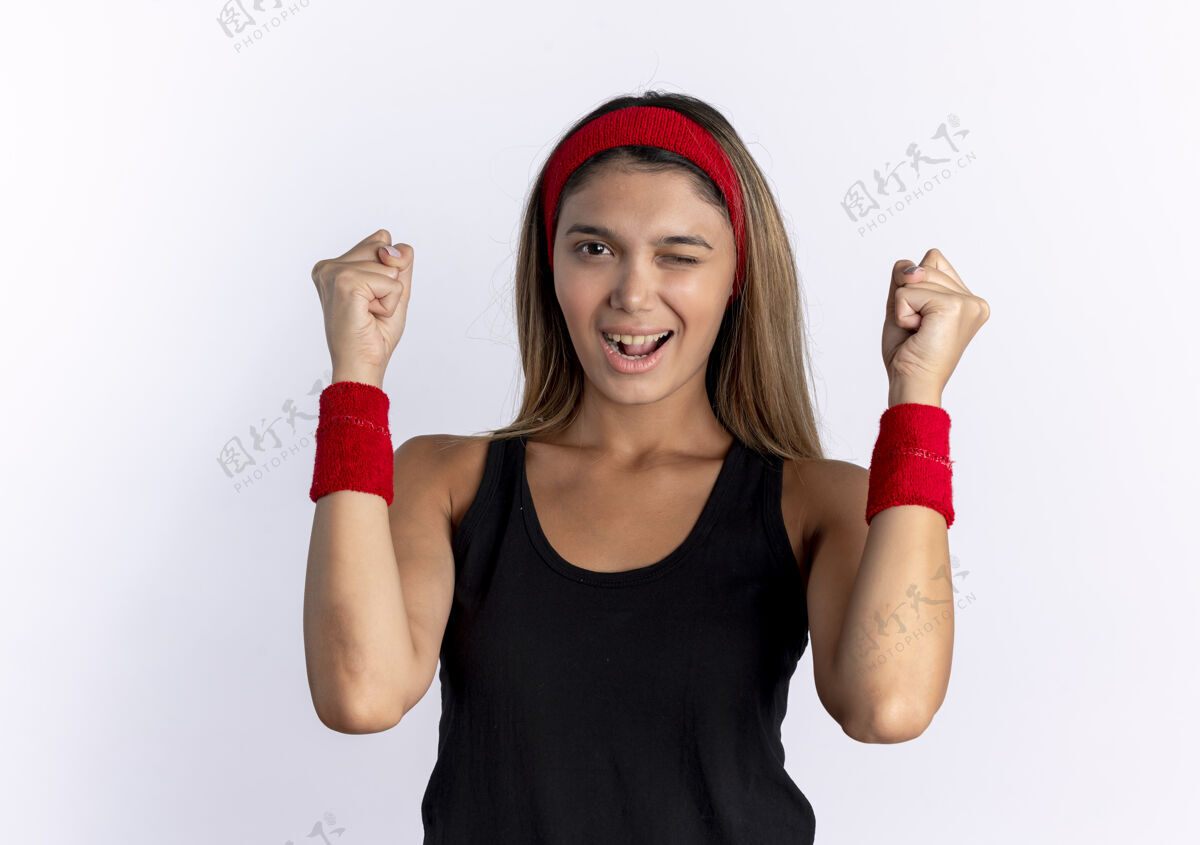 站立身穿黑色运动服 头戴红色头巾的年轻健身女孩站在白色的墙上 握紧拳头 眨眼微笑运动装女孩健身