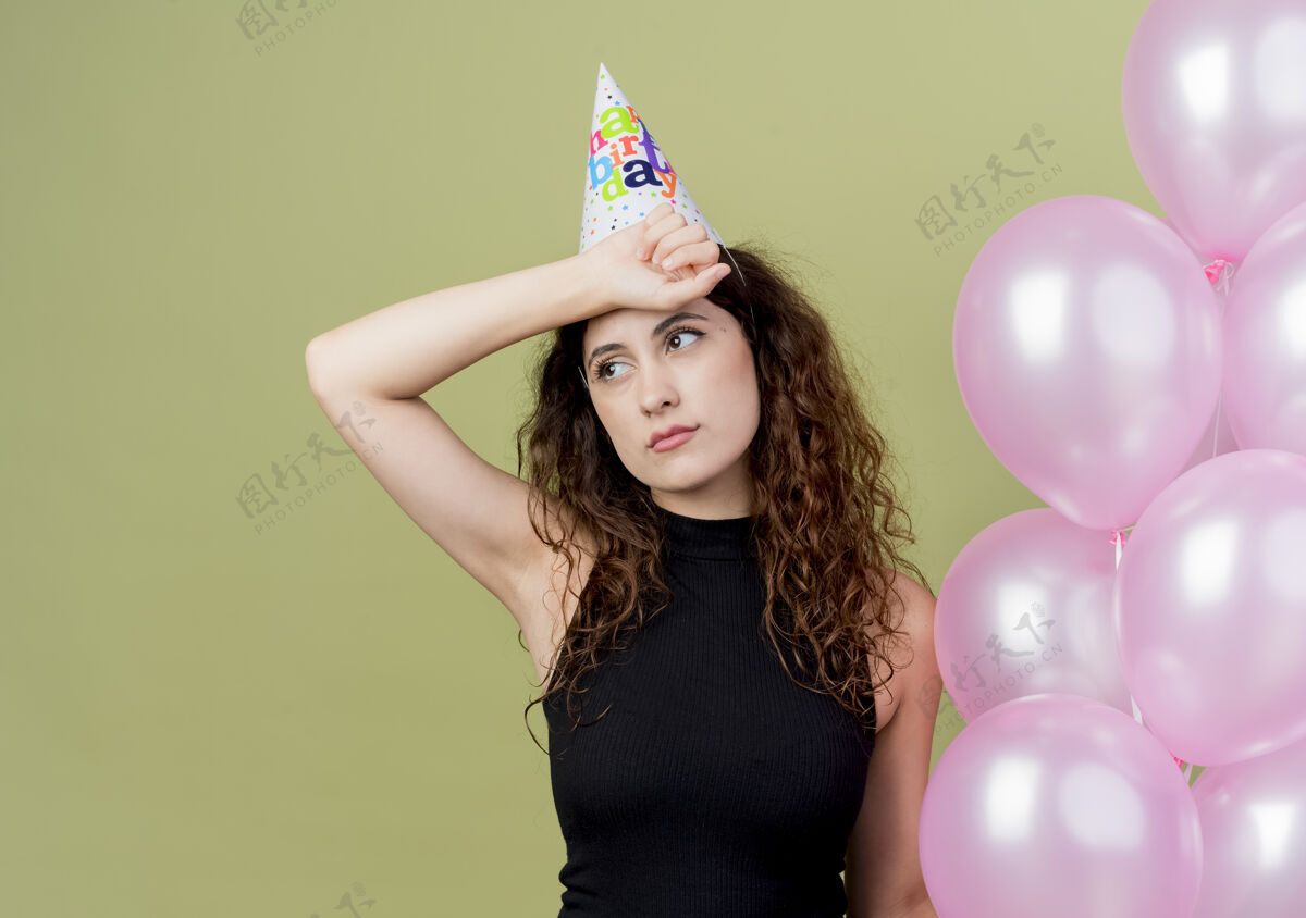 卷曲年轻漂亮的女人 卷发 戴着节日帽 手里拿着气球 看起来很累 无聊的生日派对概念站在光墙上空气光年轻