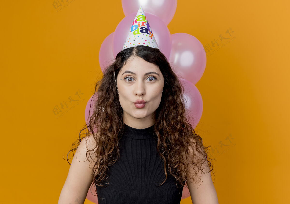 帽子戴着节日帽 留着卷发的年轻漂亮女人站在橙色的墙上看着卡内拉惊喜的生日派对概念年轻看惊喜