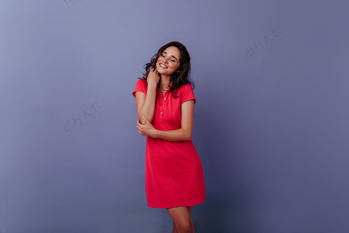 配饰迷人的年轻女子闭着眼睛微笑着站在紫色的墙上室内照片是一个穿着时髦的红色裙子的漂亮卷发女孩服饰动作魅力