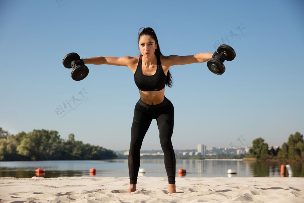 力量年轻健康的女子在沙滩上练习上身举重河流健美肌肉