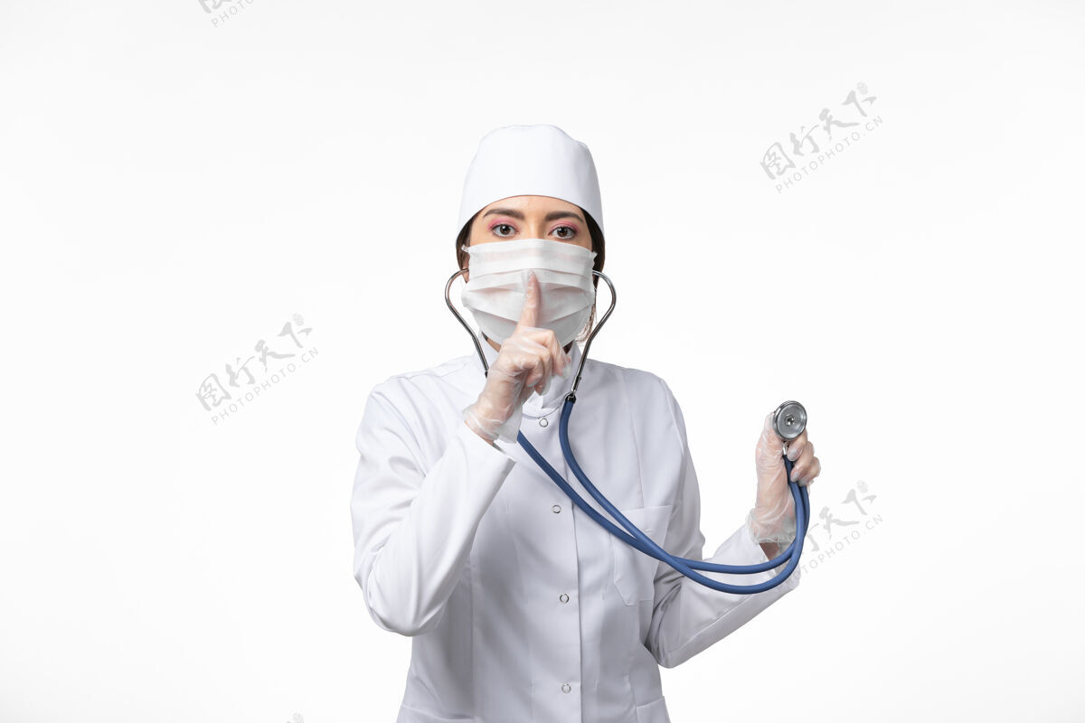 冠状病毒防护正面图女医生身穿白色无菌医疗服 戴口罩因冠状病毒-使用听诊器对白墙疾病冠状病毒-大流行性疾病冠状病毒预防视图使用