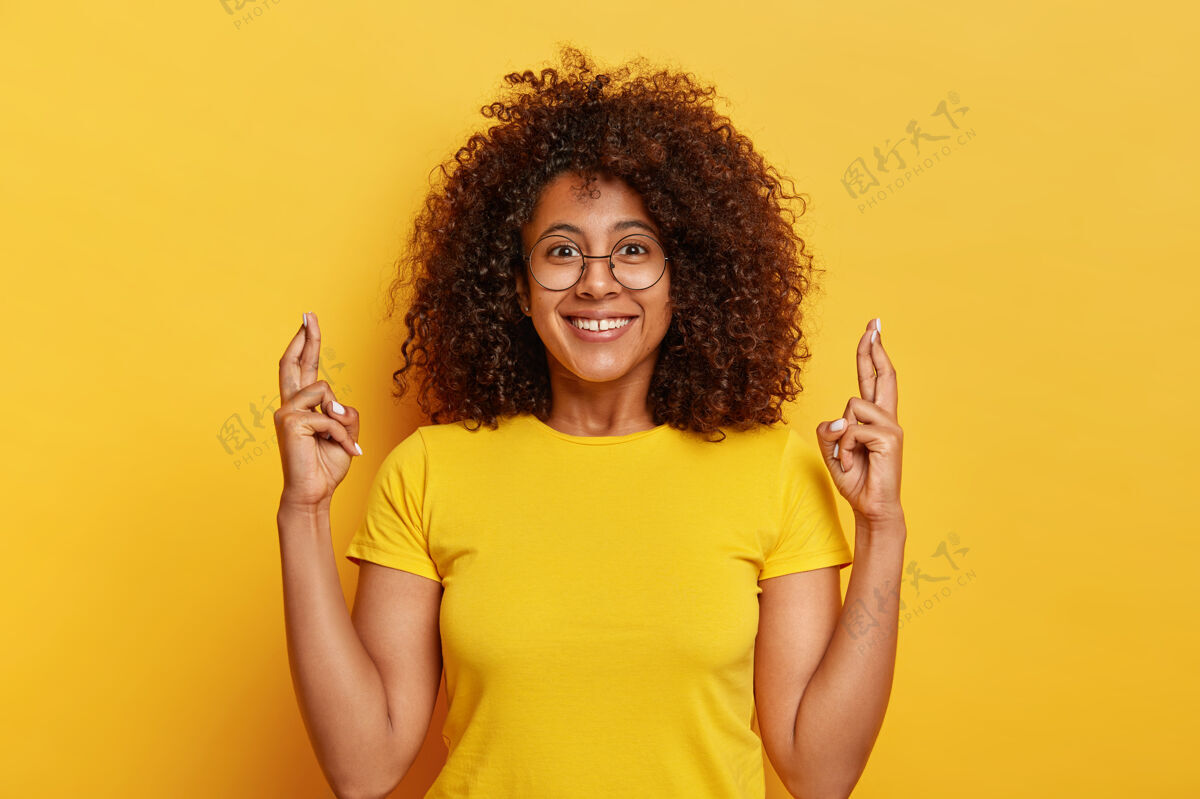 梦想穿黄色t恤的漂亮女人 交叉手指 憧憬幸运的未来 愉快地微笑 在生动的背景下摆出姿势 在室内祈祷热情表情单色