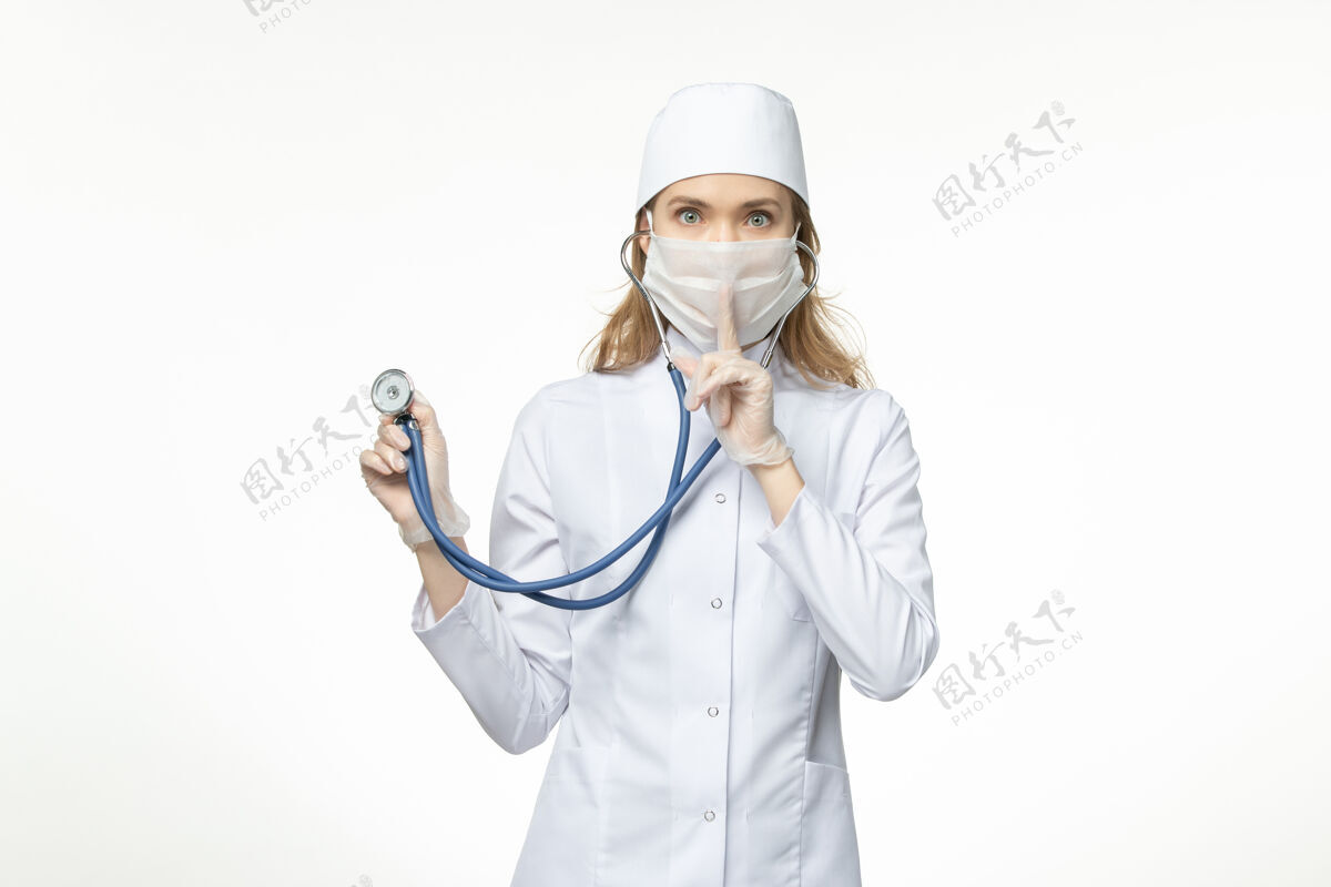 听诊器正面图女医生身穿医疗服戴口罩因冠状病毒手持听诊器在白墙上大流行冠状病毒病正面女性手持