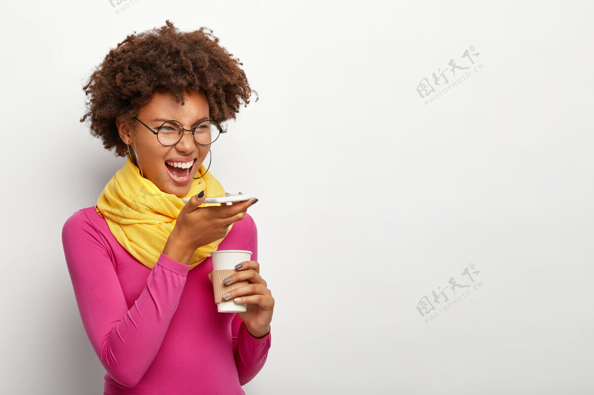 音频一个情绪化的黑皮肤卷发女人的侧面镜头 在现代手机上使用语音识别应用程序 拿着外卖咖啡 戴着眼镜 玫瑰色高领毛衣 在白墙上摆姿势电话文案空间饮料