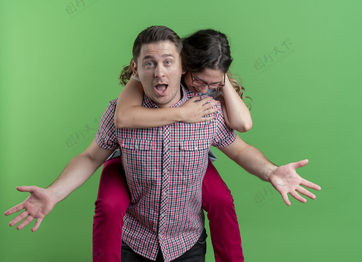 背一对年轻开朗的夫妇微笑着背着他笑着的女友站在绿色的墙上男人情侣笑