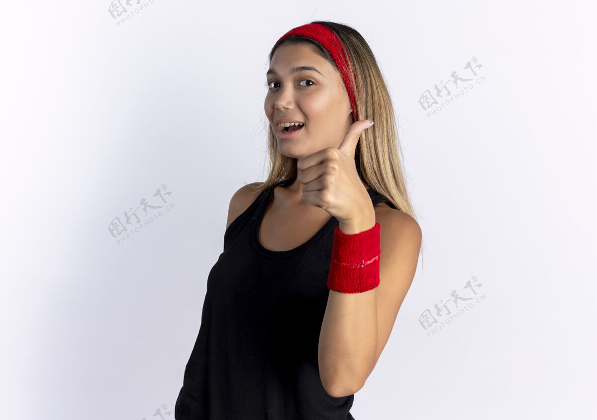 健康身穿黑色运动服 头戴红色头巾 微笑着竖起大拇指站在白墙上的年轻健身女孩年轻女孩看
