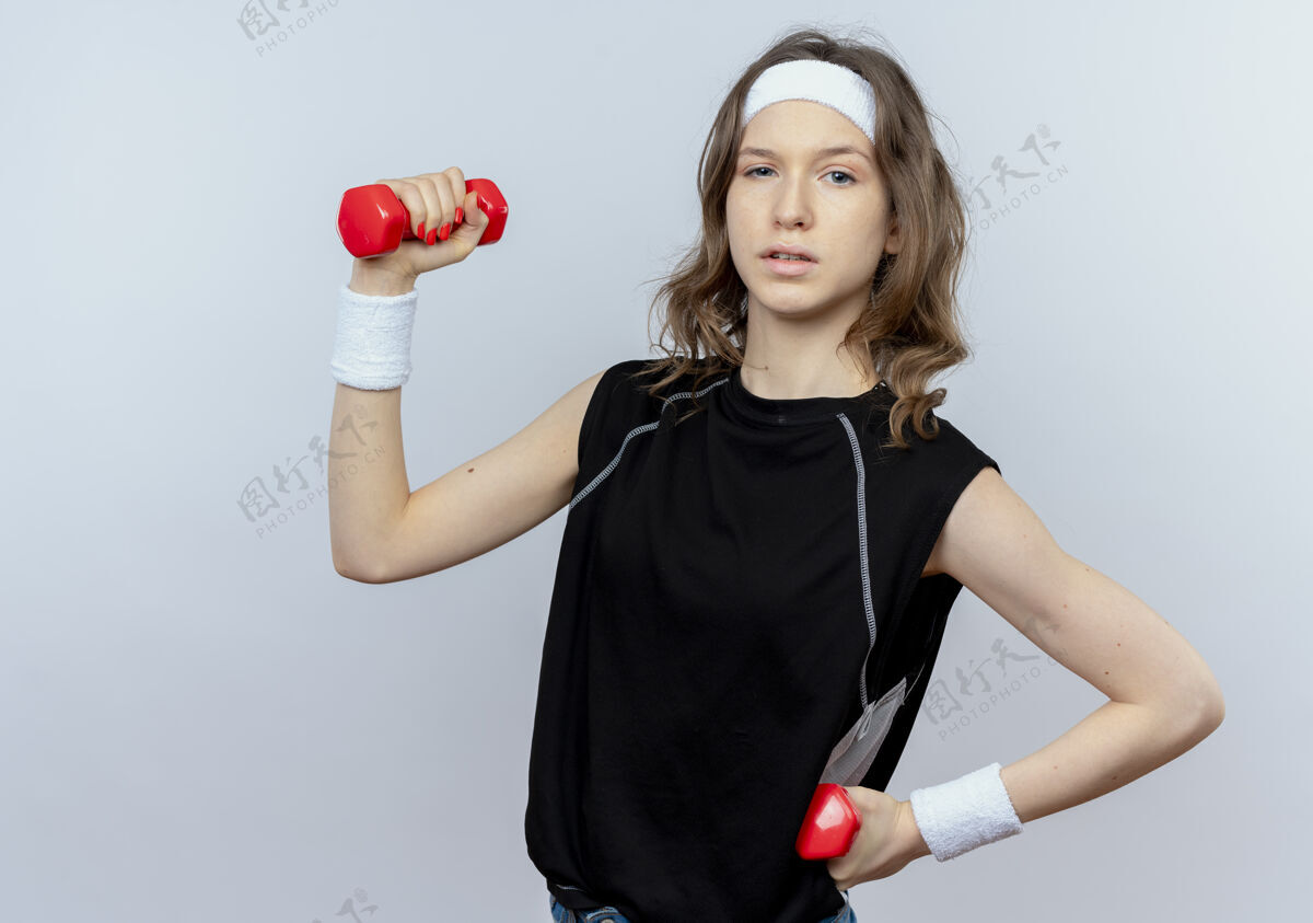 运动装身穿黑色运动服的年轻健身女孩 戴着头带 举着哑铃 站在白墙上 看起来很自信健身站立工作