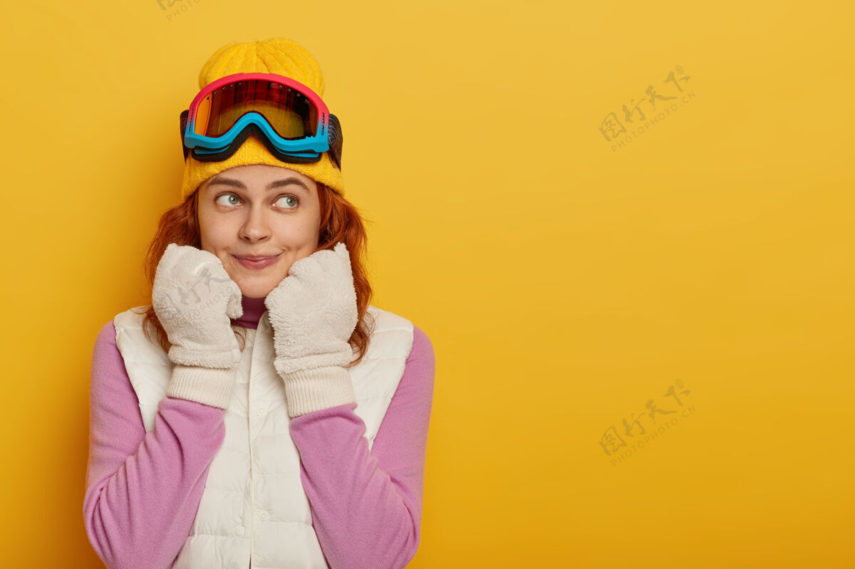 喜悦运动型的滑雪者一边沉思着 一边戴着白色的冬季手套和背心 一边戴着滑雪护目镜 一边看 一边对着黄色的工作室墙壁摆姿势 空白处白种人女人手持