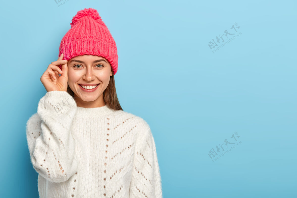 毛衣漂亮的黑发年轻女子手贴脸 微笑愉快 戴着帽子和白色针织毛衣 表达良好的情绪 在蓝色的工作室墙上摆姿势触摸天气帽子