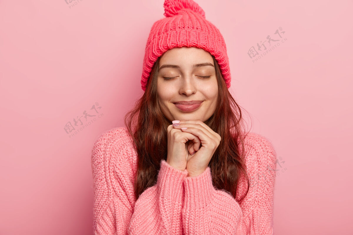满意一幅美丽迷人的女人的画像 双手放在下巴下 闭着眼睛 陷入沉思 模特们站在玫瑰色的画室墙上 穿着毛衣 戴着帽子女士冬季高兴