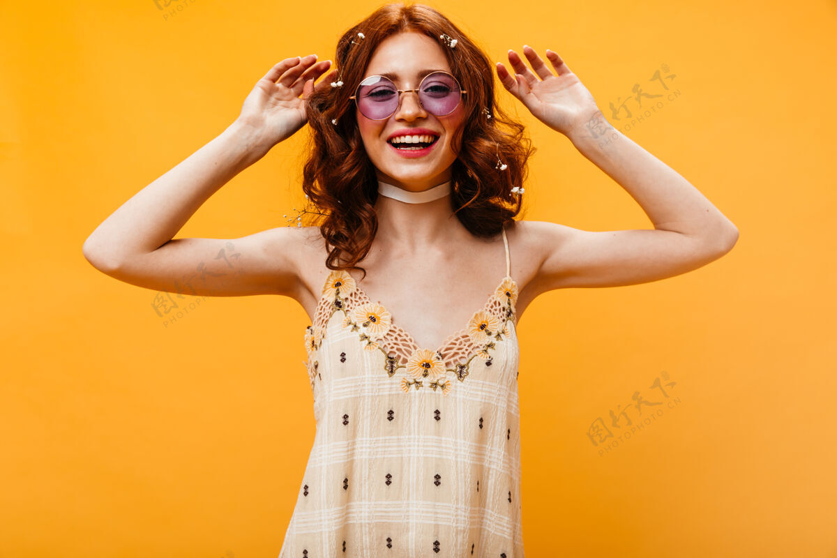 女士可爱的卷发女人 头发上插着鲜花 在橙色背景下翩翩起舞戴着眼镜和黄色太阳裙的女人的照片发型太阳裙花