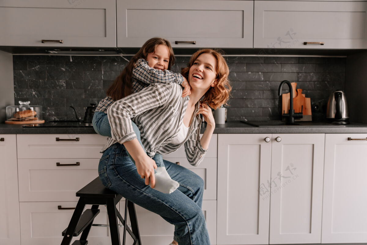 家庭穿着牛仔裤的漂亮女人坐在厨房的椅子上 她的女儿从后面拥抱着她食物妈妈糕点