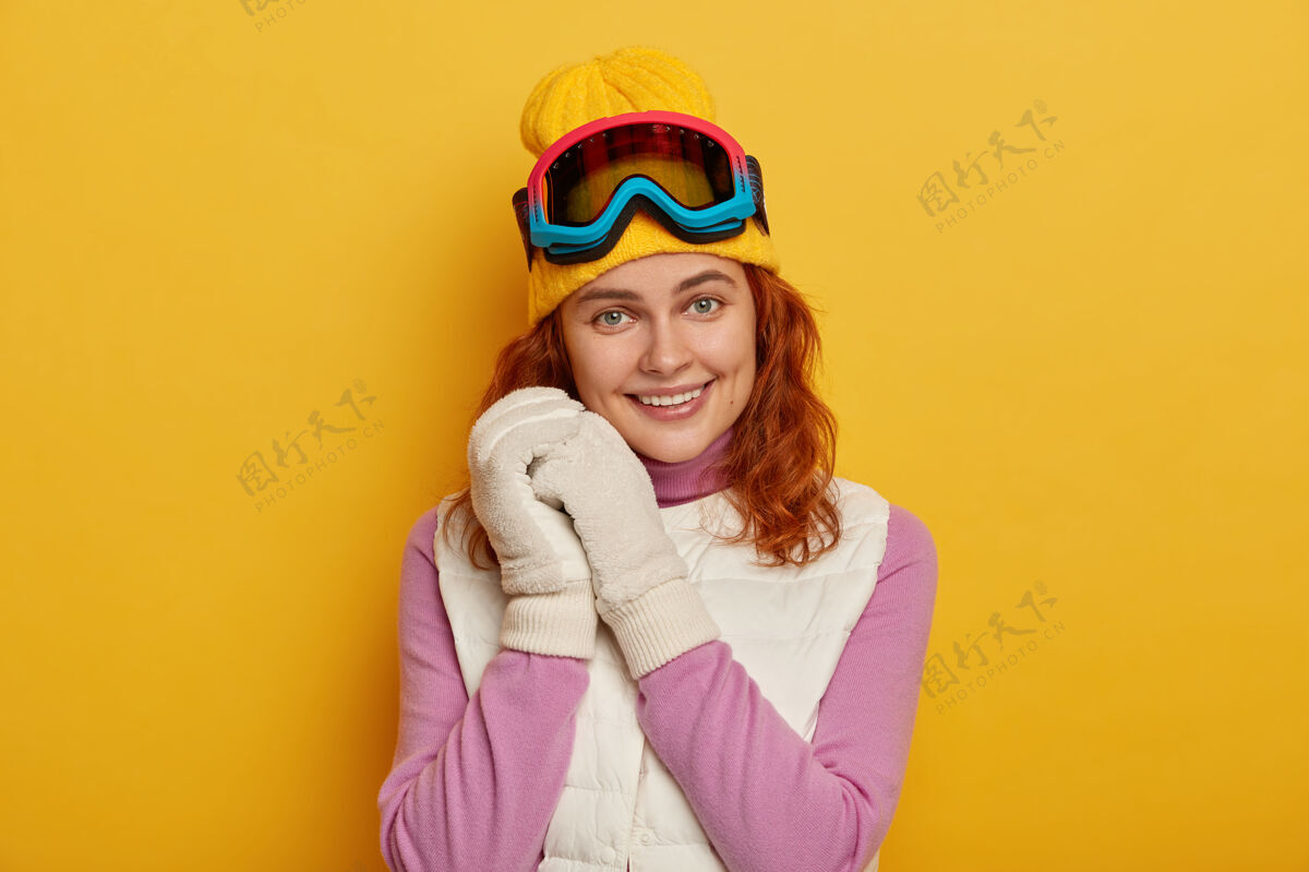背心开朗的姜女双手贴在脸上 温柔地笑着 戴着滑雪镜 穿着暖和的衣服 隔着黄色的工作室墙手套运动女人