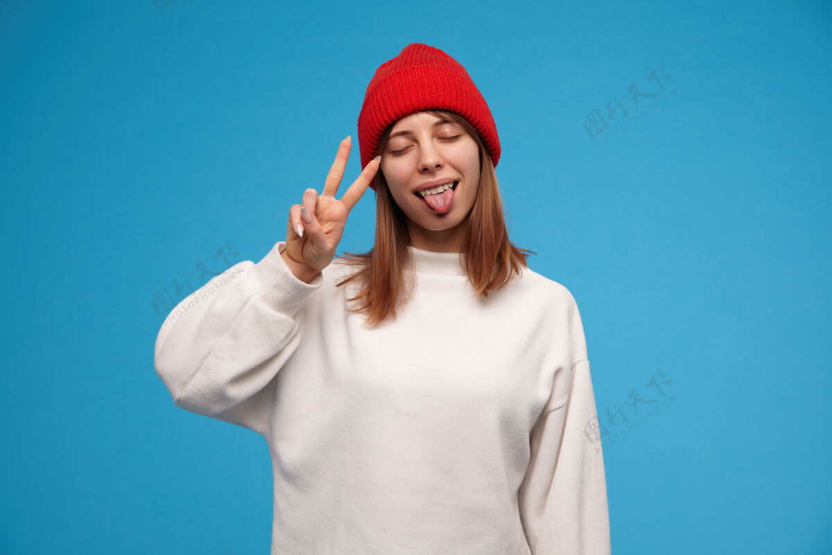 年轻有吸引力的 深色头发的成年女孩的肖像穿着白色毛衣和红色帽子显示和平标志和舌头 保持眼睛闭上站在蓝色的墙隔离服装休闲模型