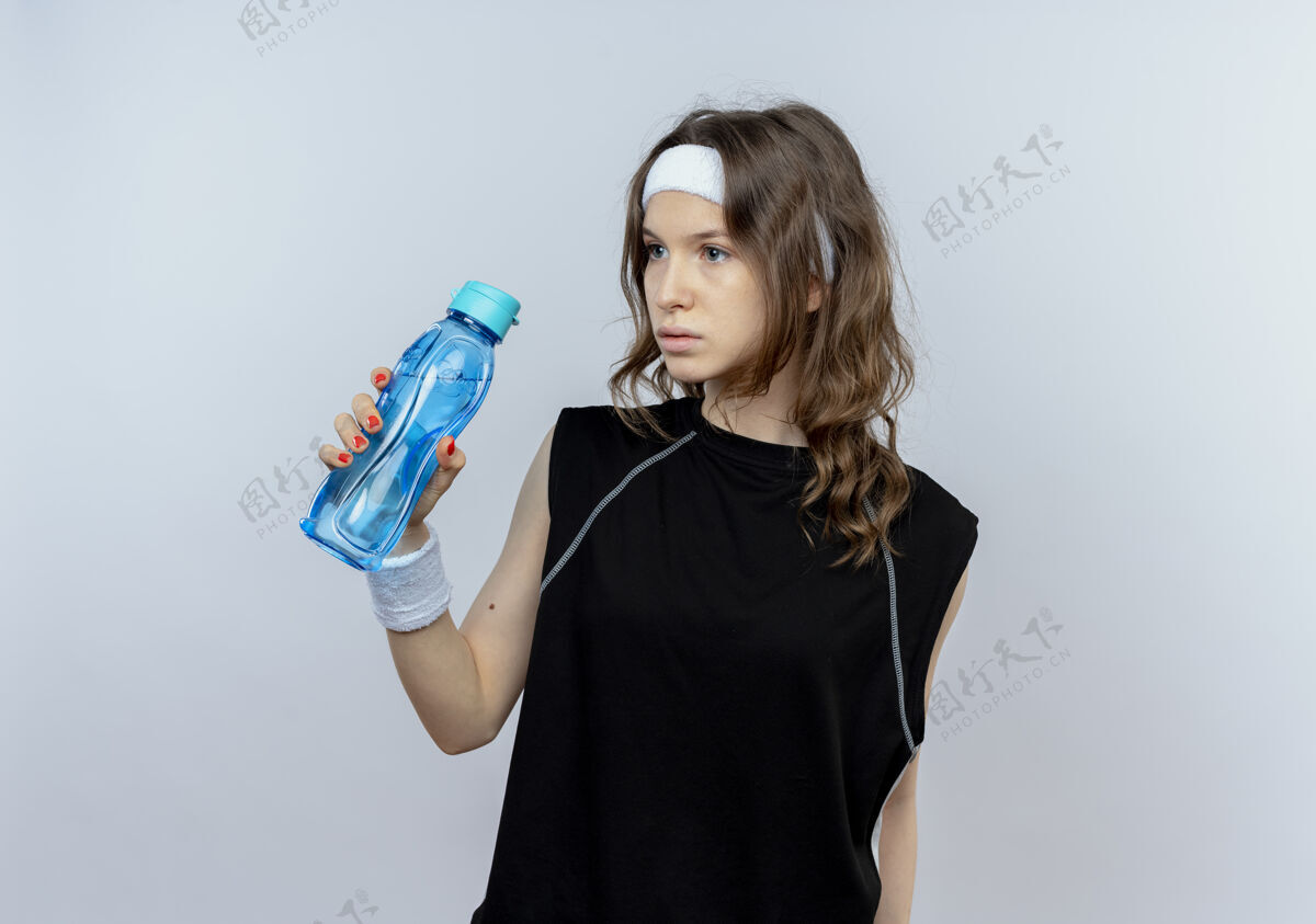 站身穿黑色运动服的年轻健身女孩戴着头巾 手里拿着一瓶水 站在白色的墙上茫然地看着一边头带健身看