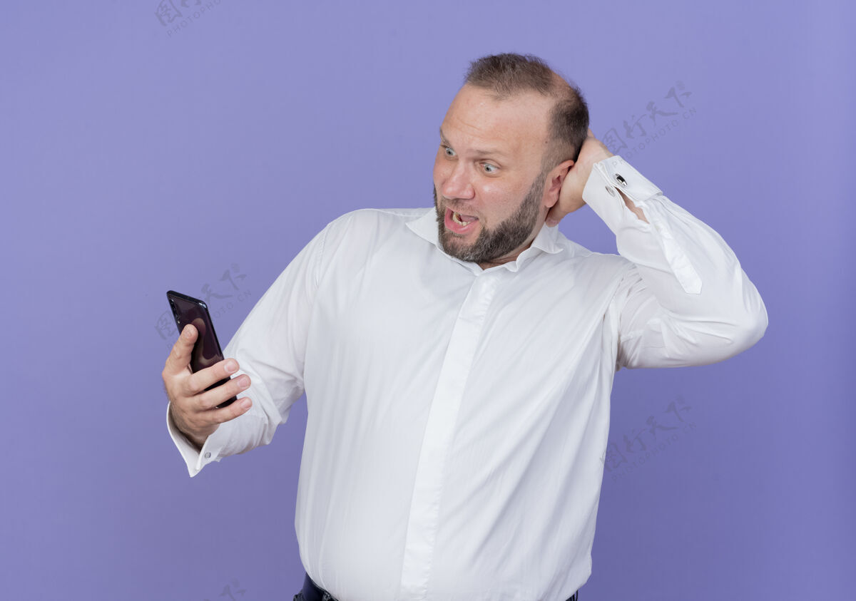 抱着的一个留着胡子的男人穿着白衬衫 手里拿着智能手机 站在蓝色的墙上 看上去既困惑又惊讶惊讶的移动的困惑的