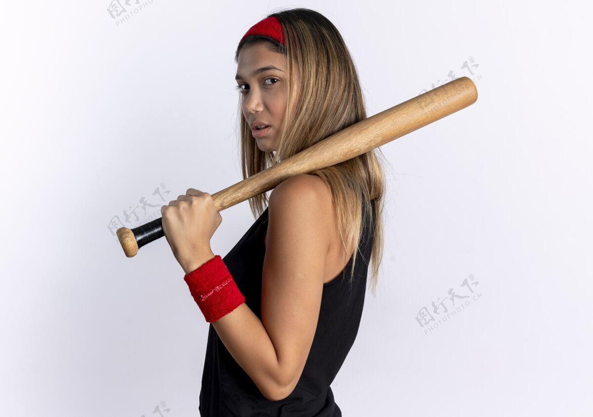 信心身穿黑色运动服 头戴红色头带 手持棒球棒的年轻健身女孩站在白色的墙上 看上去很自信看站年轻