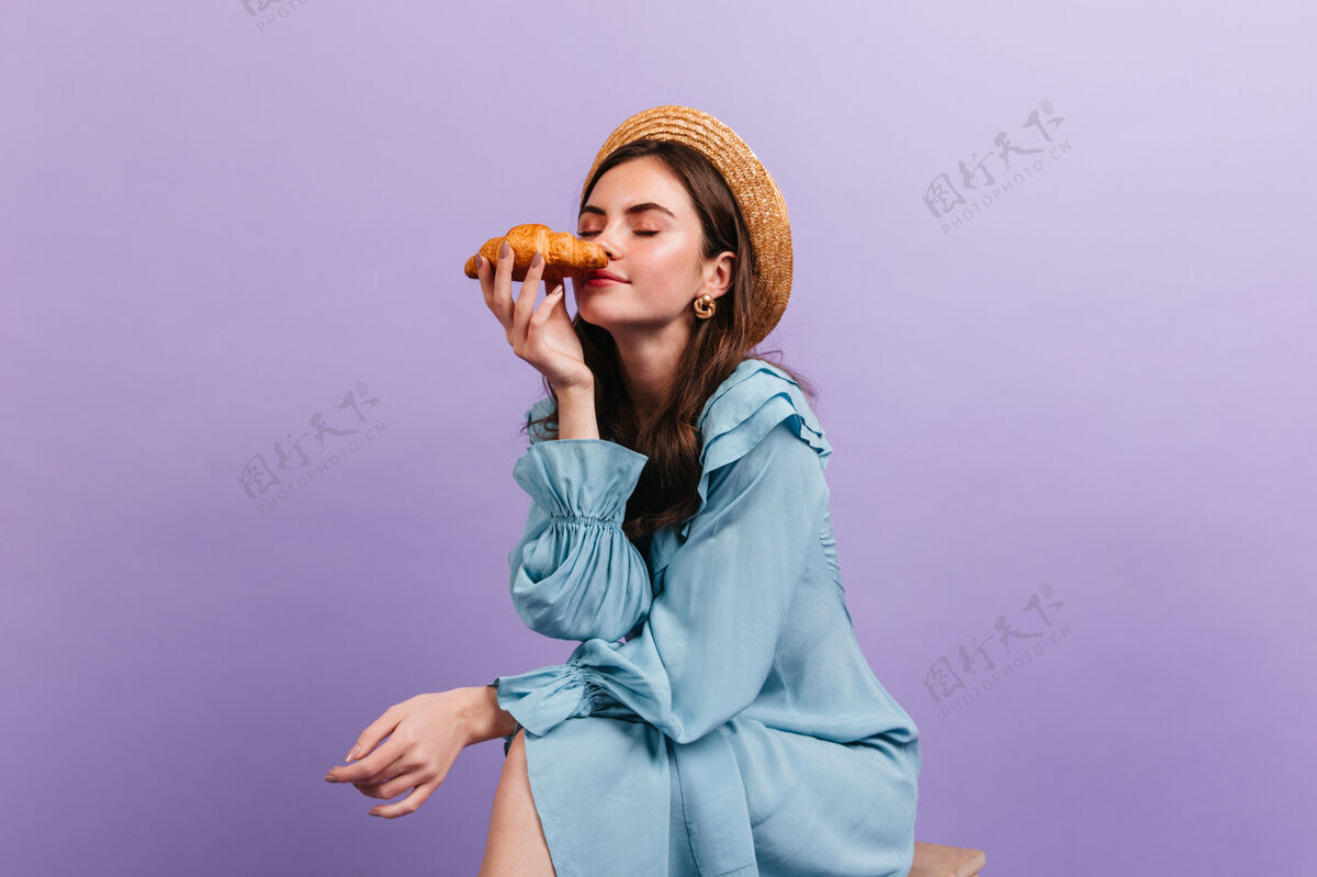 卷发一幅深黑色卷发女郎享受羊角面包香味的肖像一个穿着可爱裙子戴着帽子的女孩在紫色的墙上摆姿势微笑模特明亮