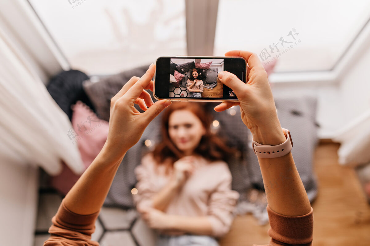 地板手持智能手机拍照的照片红发女士为朋友摆姿势的室内写真毛衣年轻房间