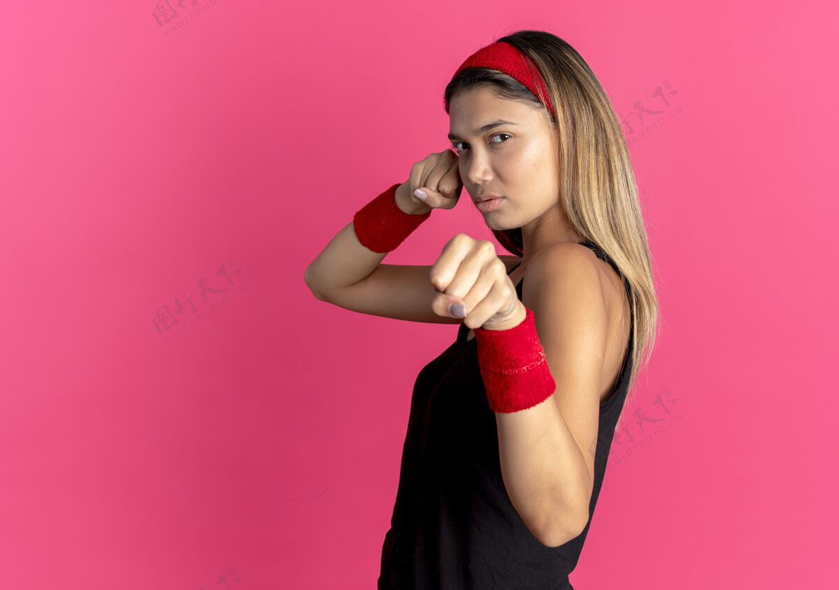 面孔身穿黑色运动服 头戴红色头巾的年轻健身女孩站在粉红色的墙上 自信地握紧拳头 表情严肃自信握紧运动装