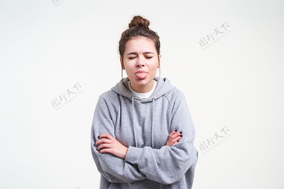 交叉室内照片 年轻的棕色头发的女性与休闲发型显示舌头闭上眼睛和折叠双手放在胸前 而在白色的背景摆姿势提高情绪白色