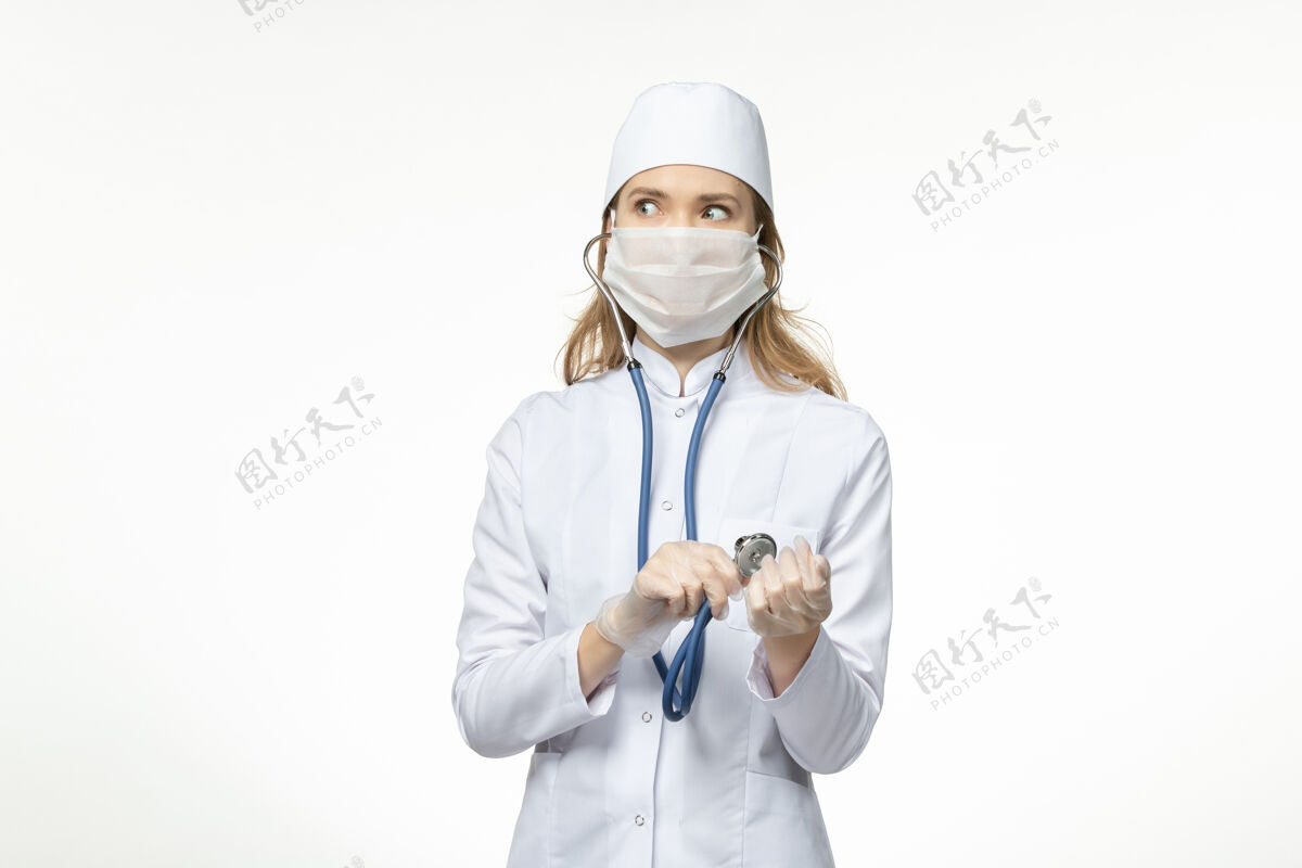 套装正面图身穿医疗服的年轻女医生因冠状病毒戴防护面罩在白色办公桌上使用听诊器冠状病毒球员办公桌