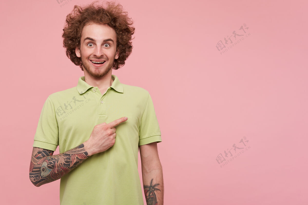 情绪年轻的卷发黑发纹身男子惊讶地圆他的绿色眼睛 同时用食指一边 在粉红色的背景隔离举高T恤马球