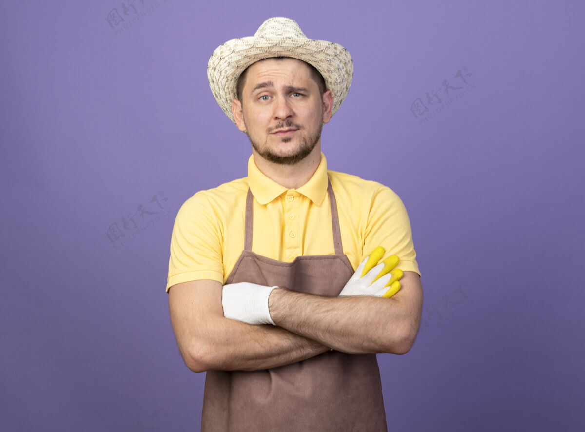 手臂年轻的园丁戴着工作手套 穿着连体衣 戴着帽子 严肃的脸 双臂交叉在胸前紫色连身衣花园