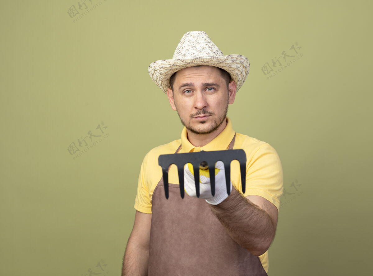 穿年轻的园丁 穿着连体衣 戴着帽子 戴着工作手套 拿着小耙子指着镜头 看上去很自信年轻人男人工作