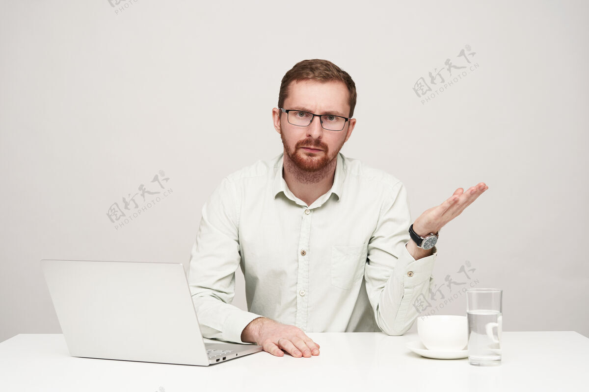 桌子恼怒的年轻人 一头金发 穿着正式的衣服 掌心抬高 眉头紧锁 严肃地看着镜头 在白色背景下摆姿势笔记本电脑坐着白色