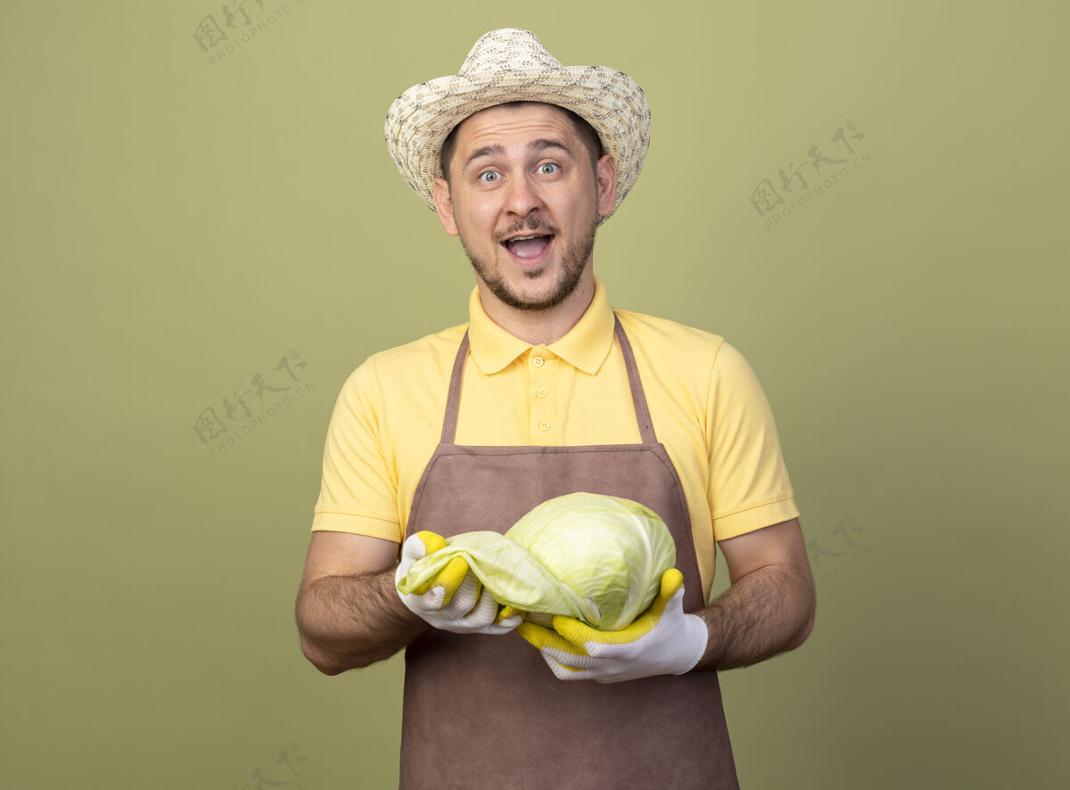 花园年轻的园丁穿着连体衣 戴着帽子 戴着工作手套 手里拿着白菜 既惊讶又高兴手套快乐卷心菜