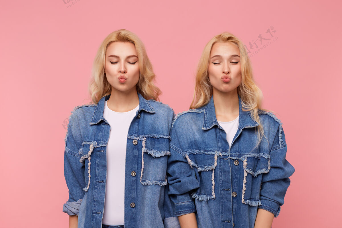 女性年轻漂亮的金发碧眼的女士 穿着牛仔裤外套和白色t恤 闭上眼睛 在粉红色的背景下 把嘴唇叠在空中亲吻表情积极双胞胎