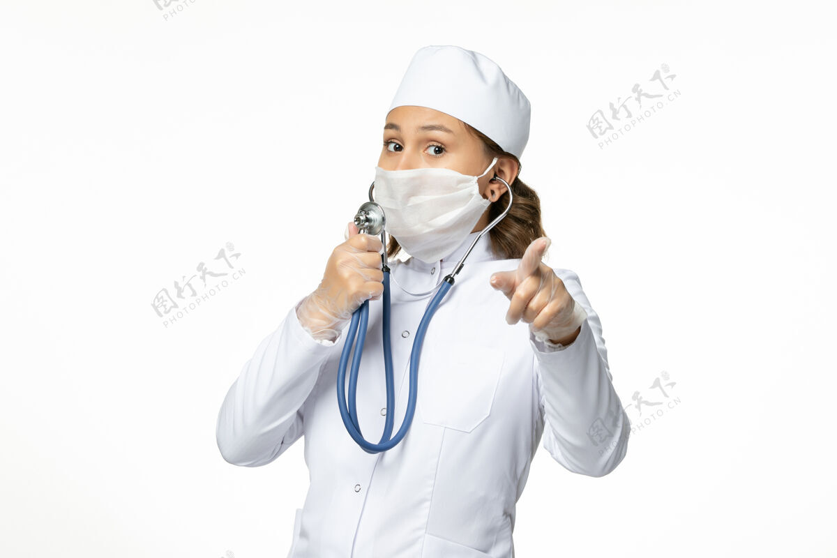 疾病正面图年轻女医生戴防护无菌口罩因冠状病毒使用听诊器在白色办公桌上大流行专业人员使用