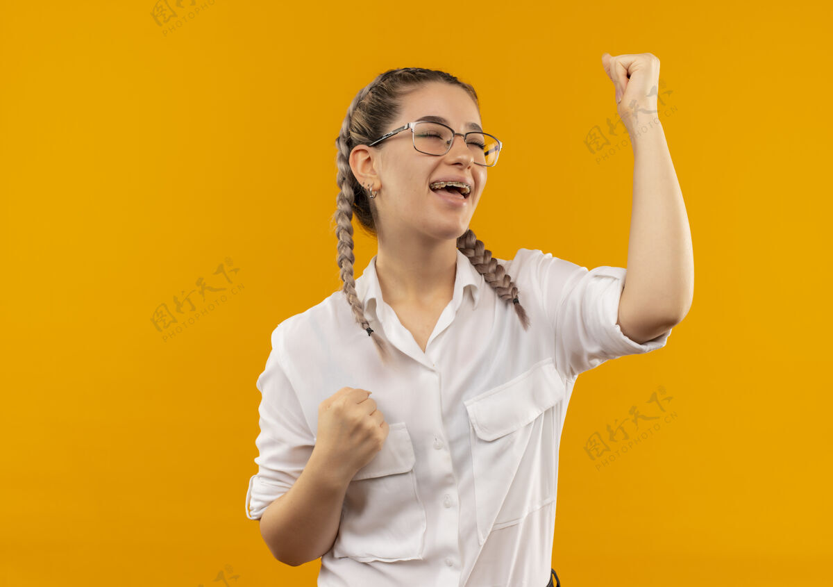 握紧戴着眼镜 梳着辫子 穿着白衬衫 握紧拳头的年轻女学生站在橙色的背景下为自己的成功而高兴和兴奋兴奋辫子白色