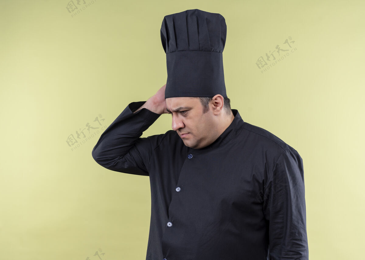 烹饪男厨师长身穿黑色制服 头戴厨师帽 困惑地看着一旁 忘记了站在绿色背景上的东西看忘了厨师