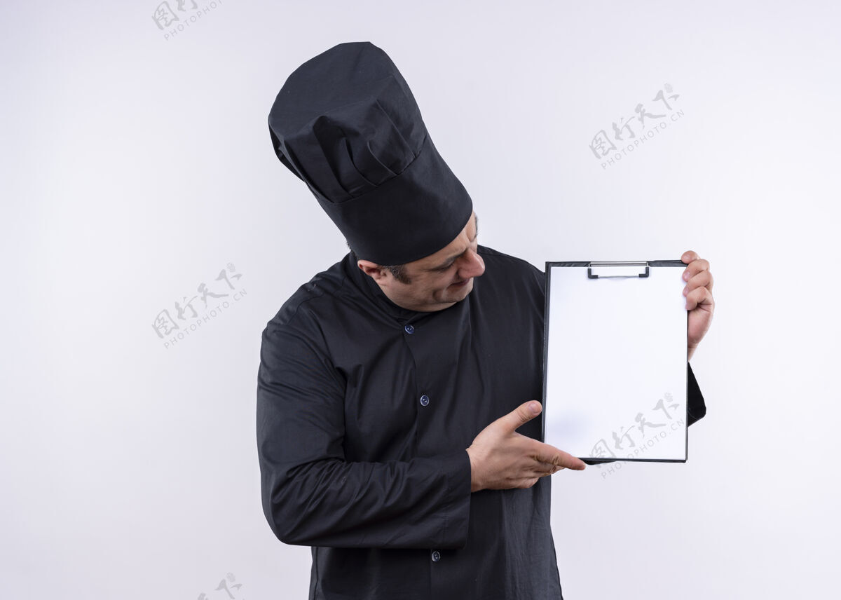 白男厨师身穿黑色制服 头戴厨师帽 拿着空白页的剪贴板 站在白色背景上看着它烹饪黑穿